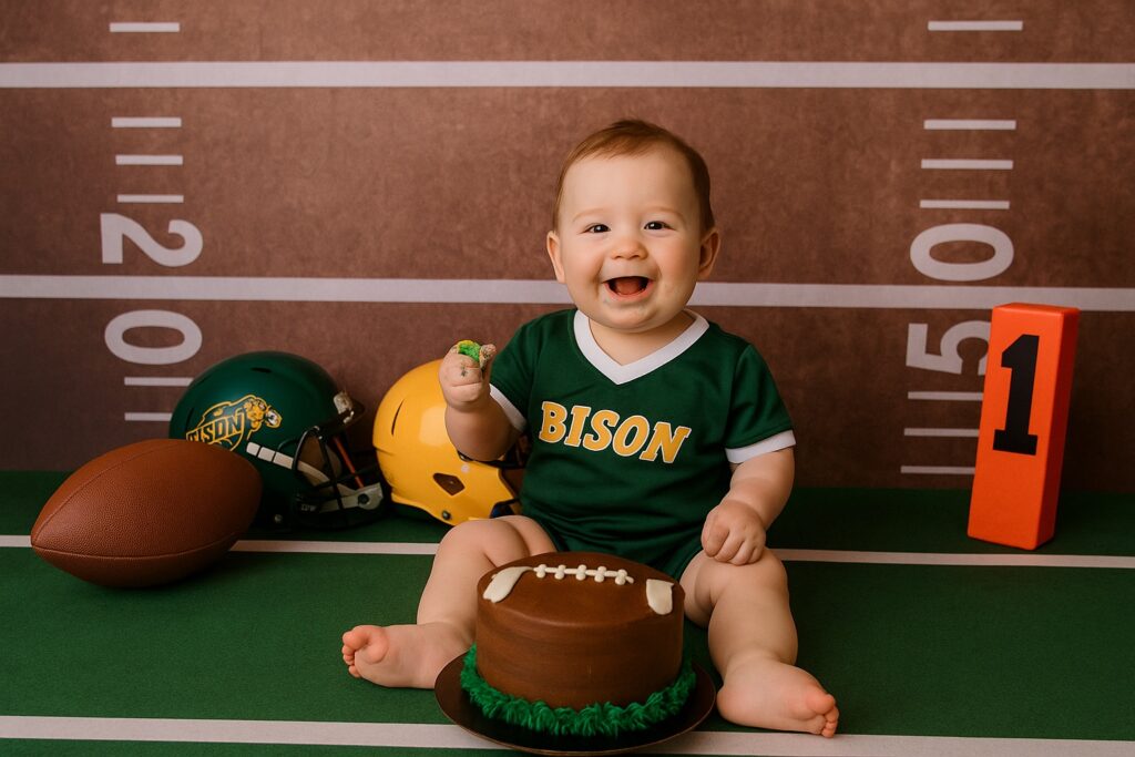 Baby boy in a green Bison jersey holding cake with football props, helmets, and a turf-style backdrop.