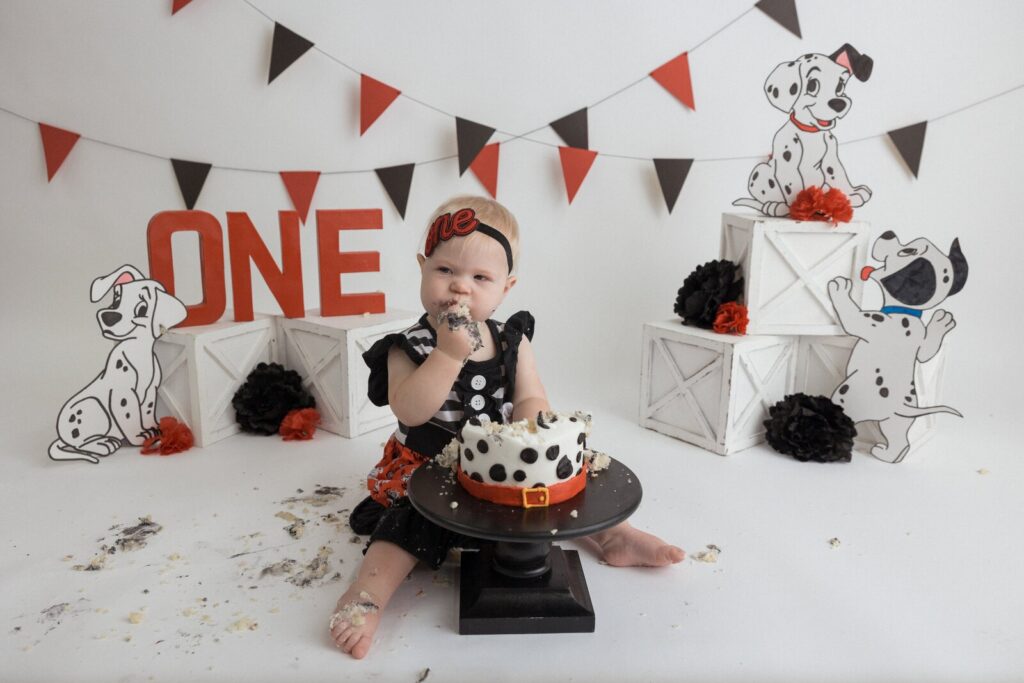 Baby boy in red and white surrounded by Dalmatian spots and a dog-themed cake, with cartoon cutouts and paw-print balloons.