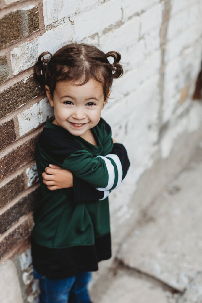 Young child with pigtails wearing a green and black jersey smiling and leaning against a white brick wall.