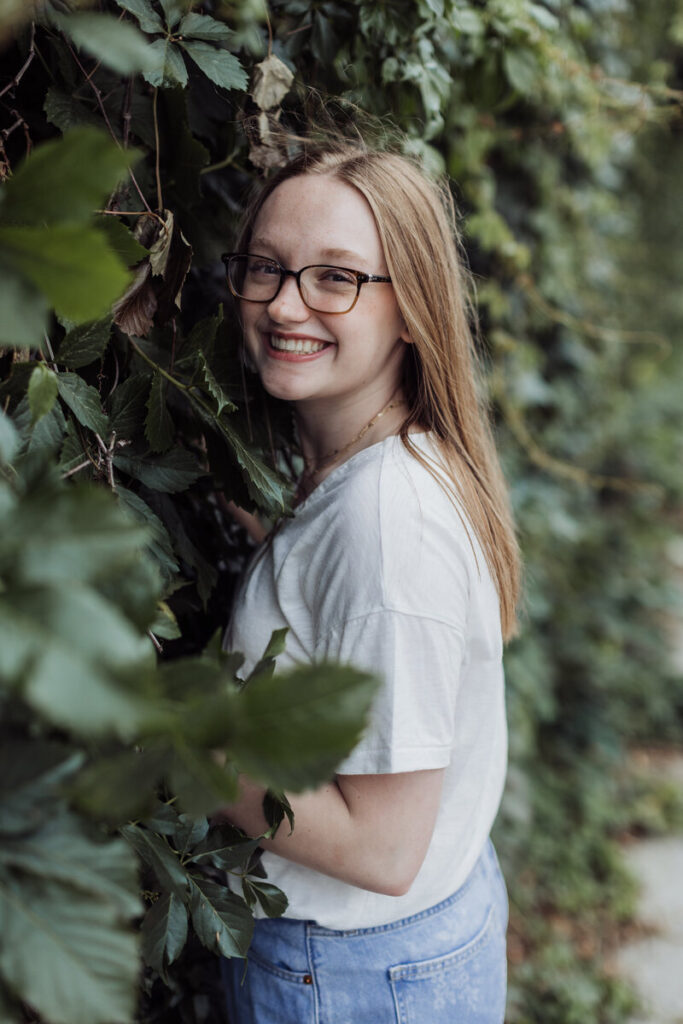 Teen girl with long hair and glasses smiling beside a leafy ivy-covered wall during a natural outdoor photo session.