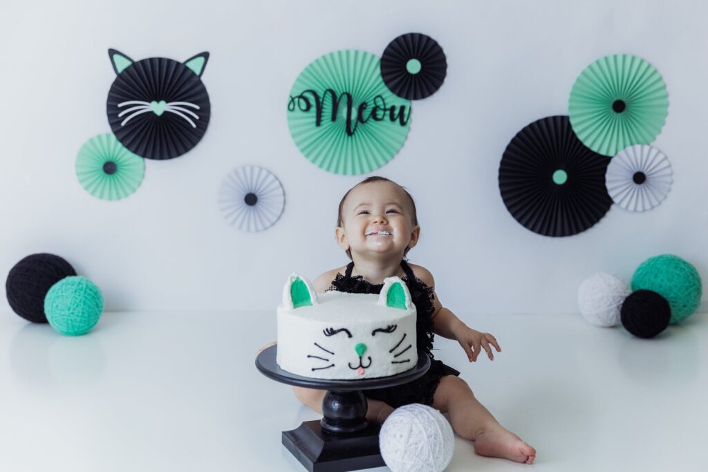 Baby girl in mint and black decor with a cat-face cake, surrounded by playful paper fans and whisker-themed props.