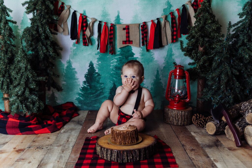 Baby boy in a red flannel shirt surrounded by pine trees and a rustic woodland setup, joyfully smashing a log-slice cake.