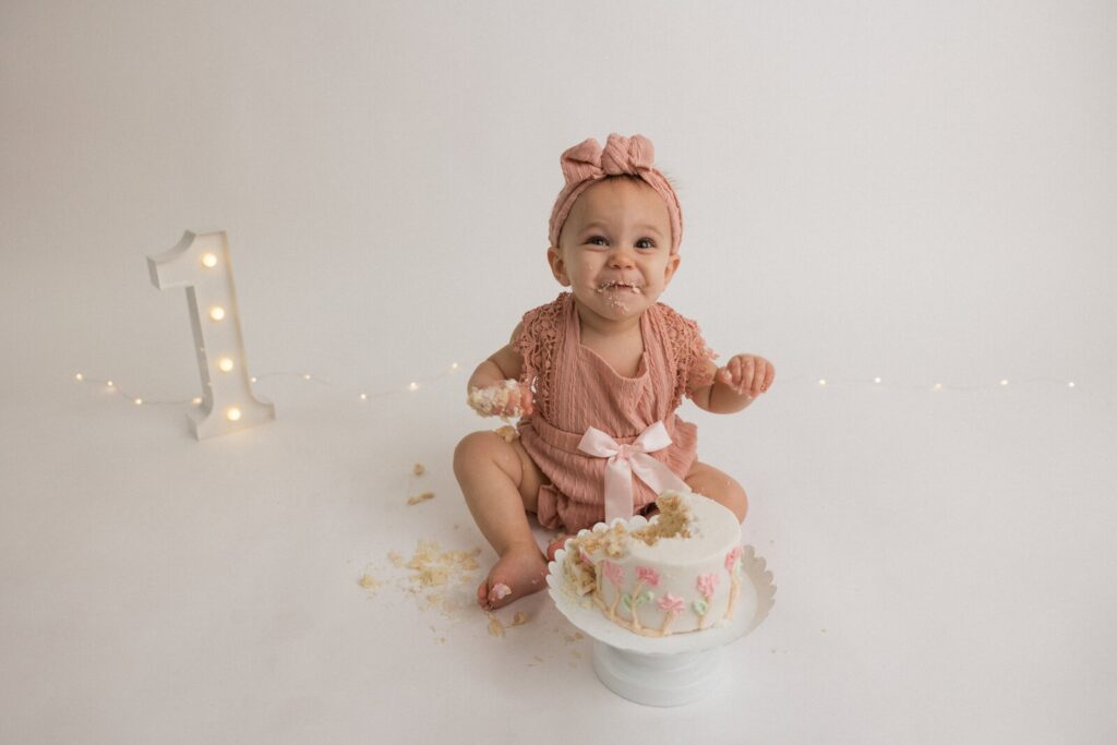 Baby girl in a soft pink romper sitting in front of a floral cake with subtle garden-themed decor in a minimalist setting.