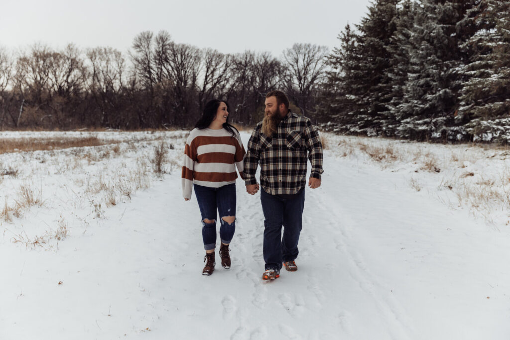 Couple holding hands and walking through the snow on a wooded trail at Forest River Park during a winter photo session.