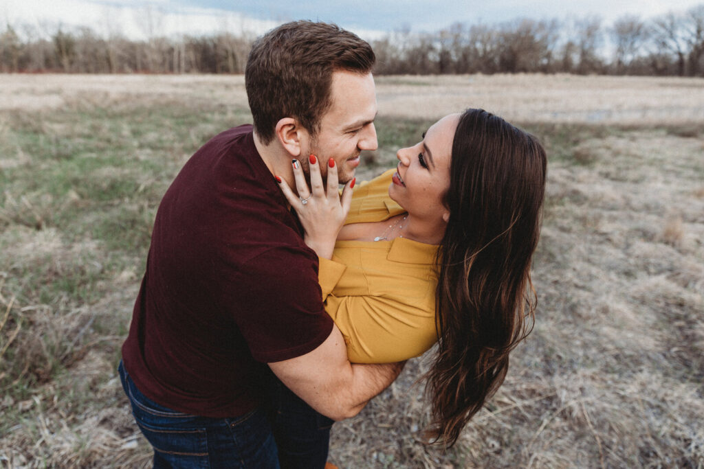 Man lifting woman on a sunlit forest path at Forest River Park in Fargo, during a golden hour engagement photoshoot.