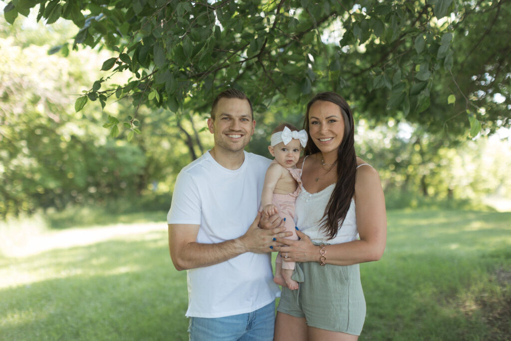 Family of three posing under leafy green trees during a sunny outdoor session at Lindenwood Park.