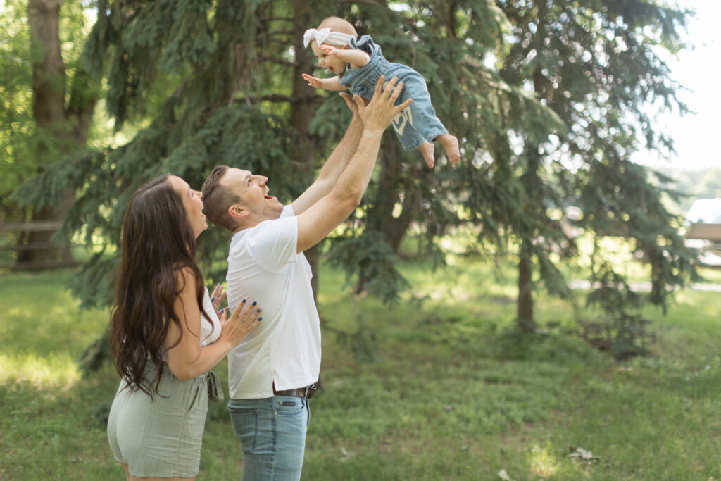 Father lifting baby girl into the air while mother smiles during a summer family photo session.