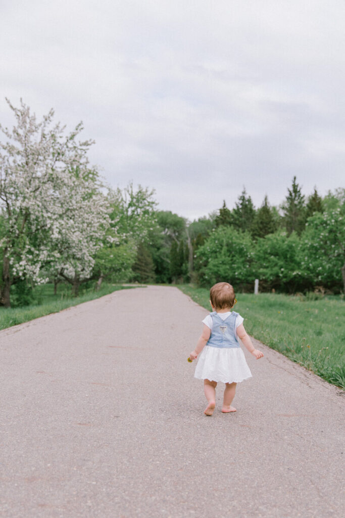Toddler in a white dress and denim vest walking barefoot along a paved path surrounded by spring trees in Orchard Glen Park.