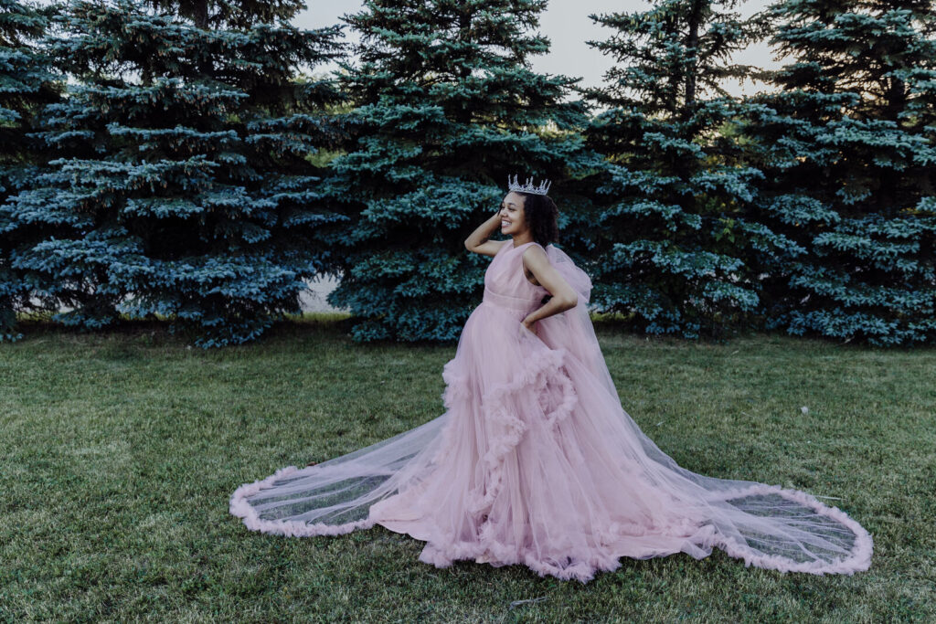 Pregnant woman wearing a flowing pink gown and crown, posing in front of evergreen trees during a maternity photo shoot at Riverwood Park