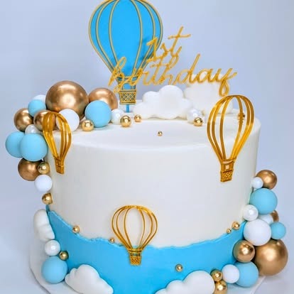A pastel blue and white cake decorated with golden and blue hot air balloons for a first birthday celebration.