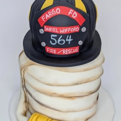 A firefighter-themed cake featuring a fondant fire helmet and hose labeled “Fargo FD.”