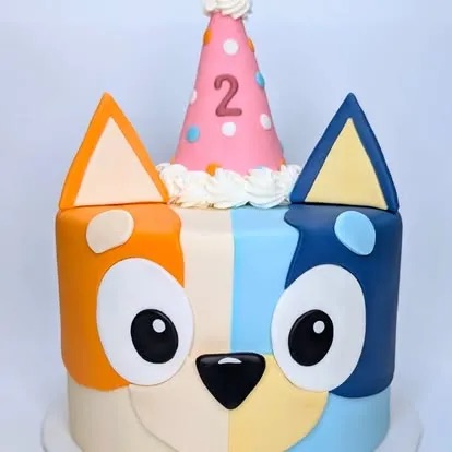 A two-tone Bluey-themed birthday cake with fondant ears and a pink polka-dot party hat featuring the number 2.