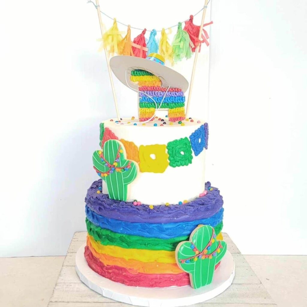 A colorful rainbow fiesta cake decorated with fondant cacti, a piñata, tassels, and bright piping.