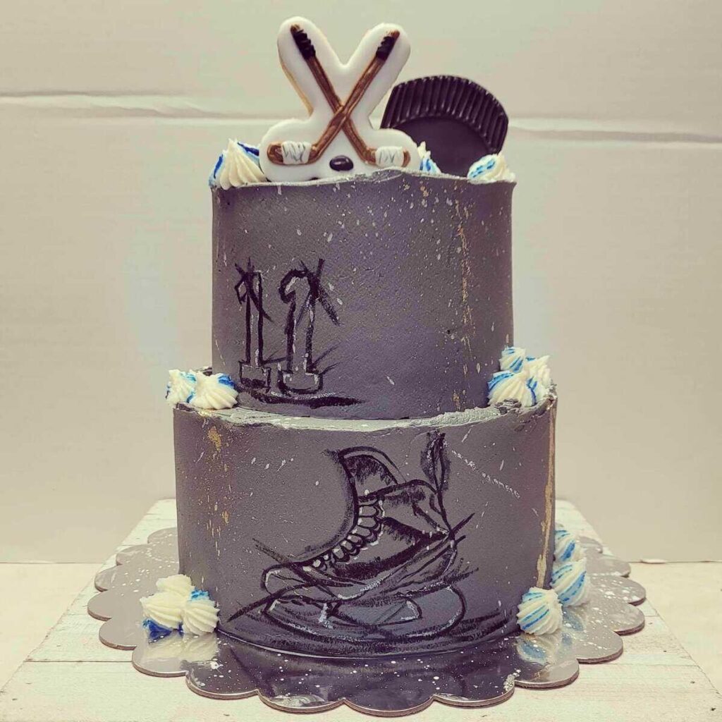 A dark gray hockey-themed cake with hand-drawn skate and number illustrations, topped with fondant hockey sticks.