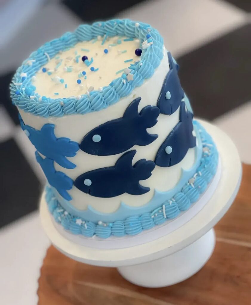 A white and blue cake with fondant fish and piped waves, topped with sprinkles.