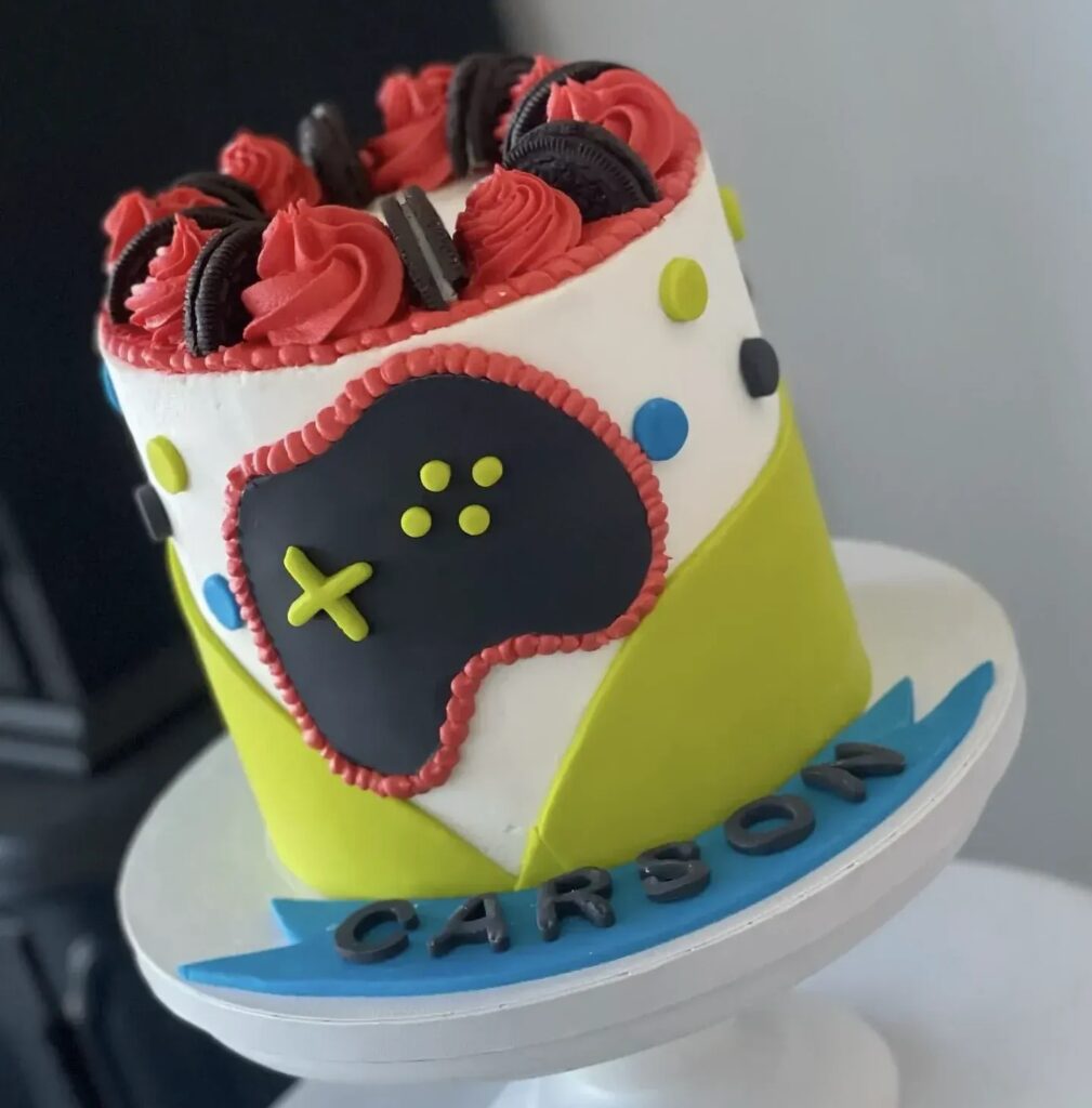 A gamer-themed cake with a fondant game controller, colorful buttons, and Oreos on top, labeled "Carson."