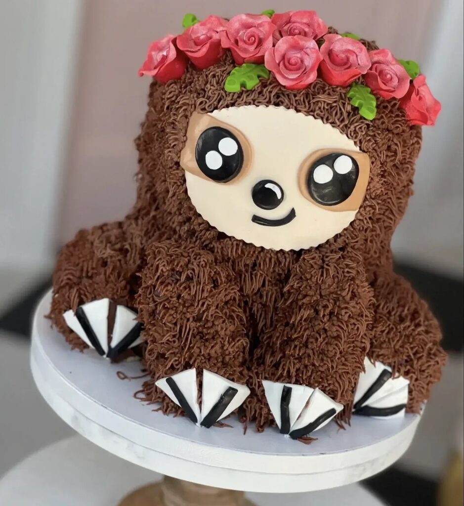 A 3D sloth-shaped cake with textured chocolate fur, a flower crown, and a cute face.