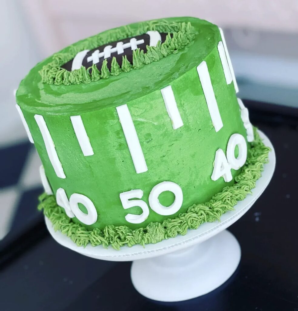 A green cake designed to look like a football field with white yard lines and a fondant football on top.