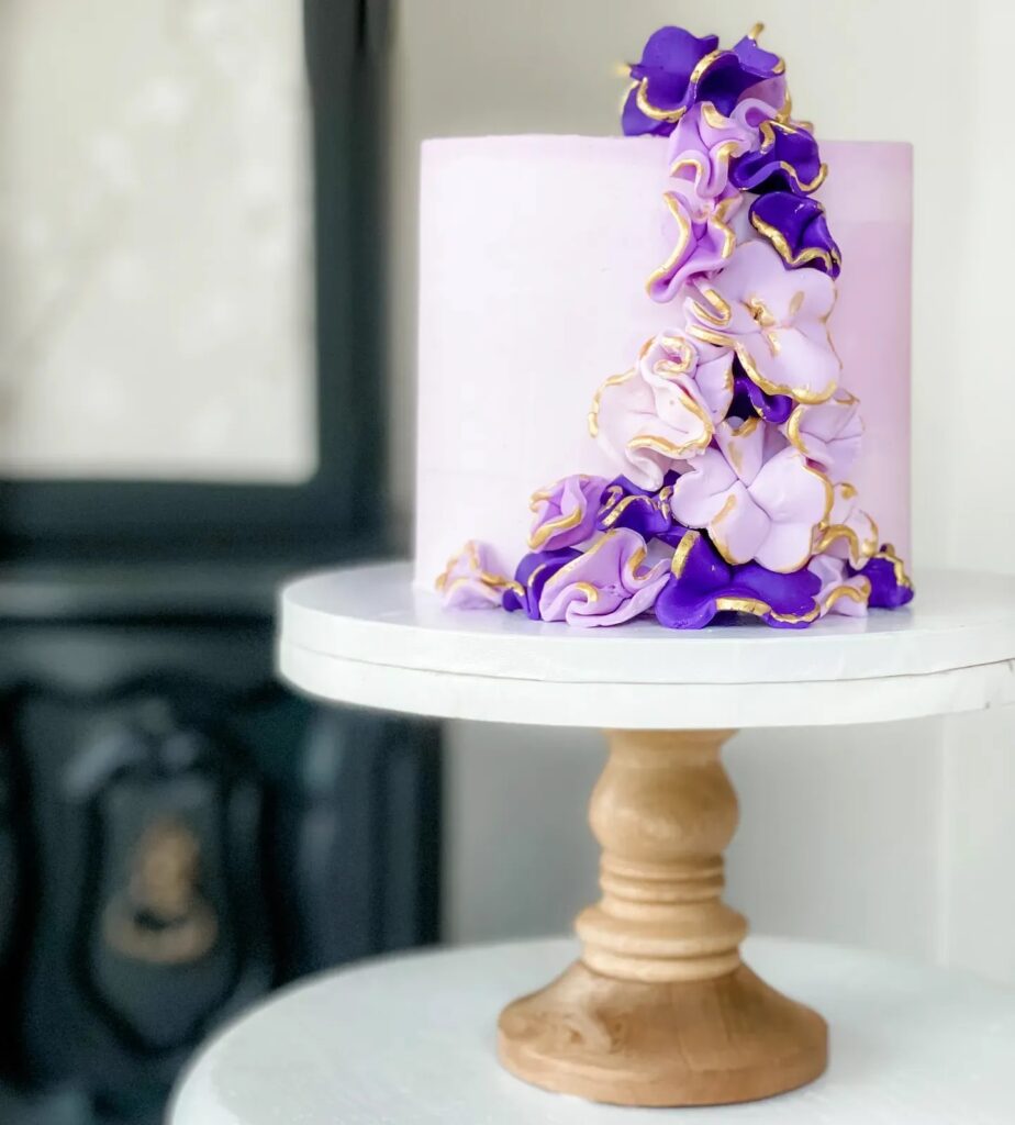 A soft pink cake with a cascading trail of purple and gold ruffled flowers.


