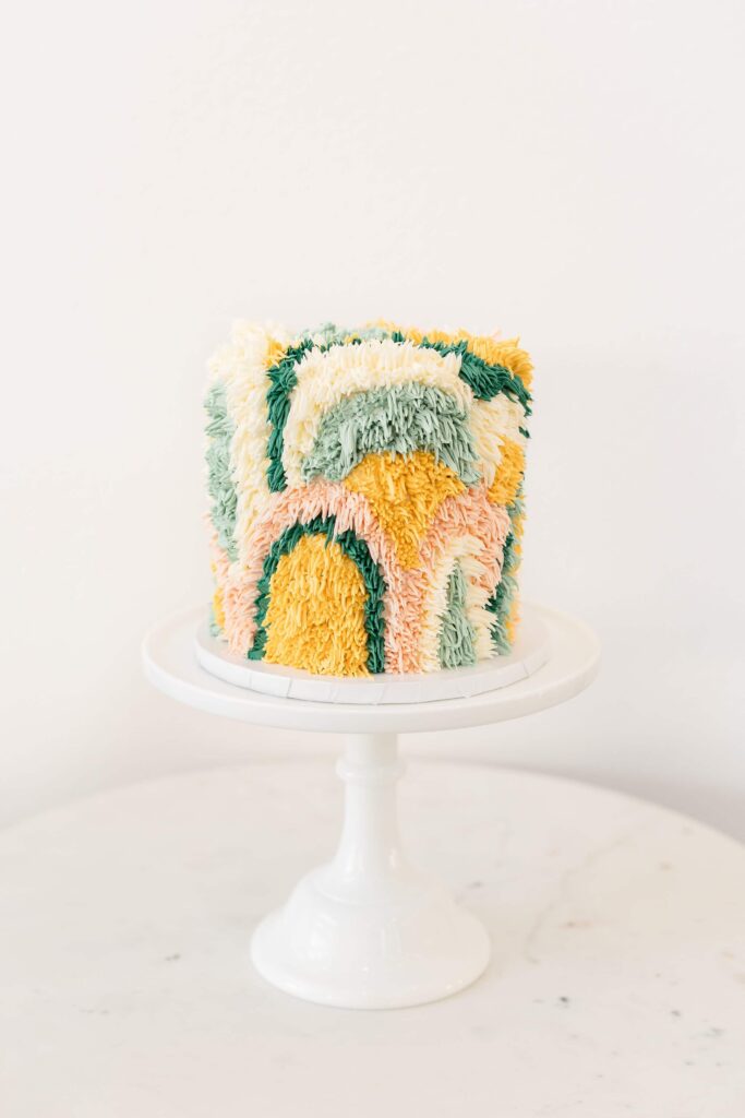 A pastel rainbow shag-textured cake with piped buttercream in earthy tones of yellow, pink, green, and white.