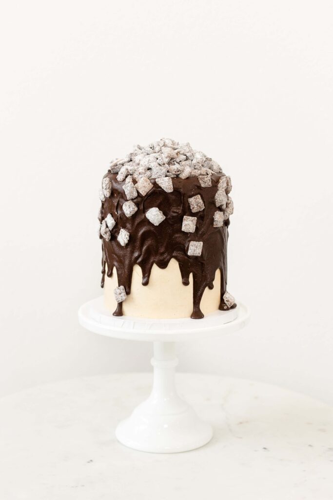 A tall cake with chocolate ganache drip, topped with powdered sugar-covered puppy chow treats.