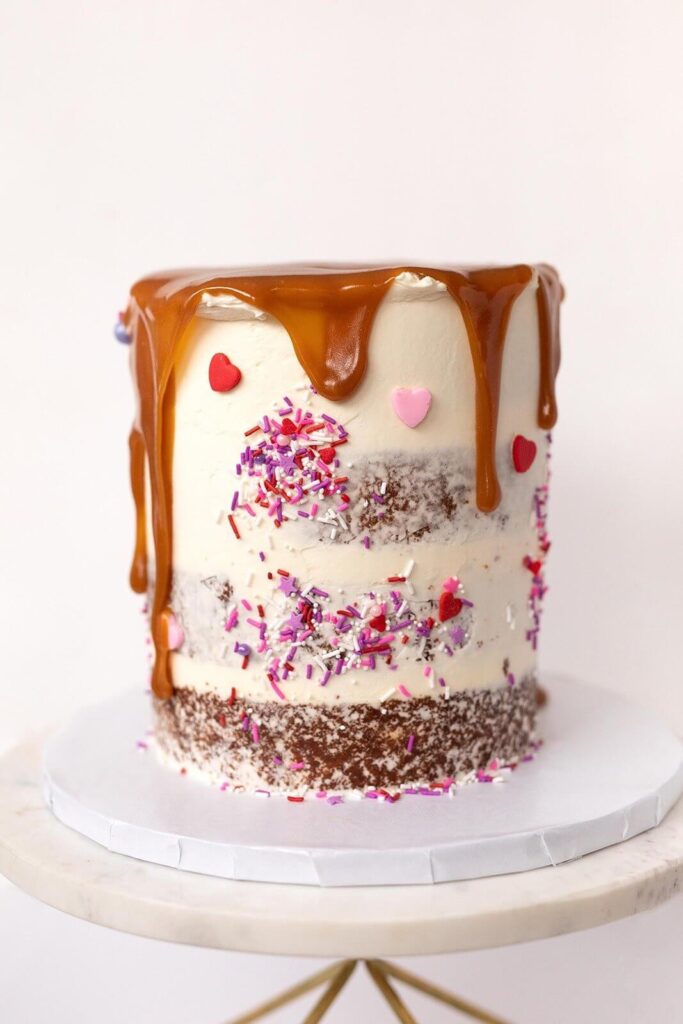 A semi-naked cake with caramel drip, heart sprinkles, and pink, purple, and red Valentine’s Day decorations.