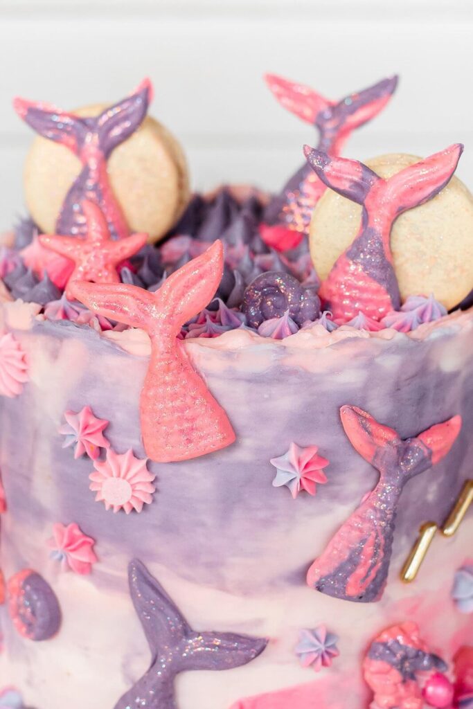 A purple and pink mermaid-themed cake decorated with glittery fondant tails, seashells, and swirls of icing.