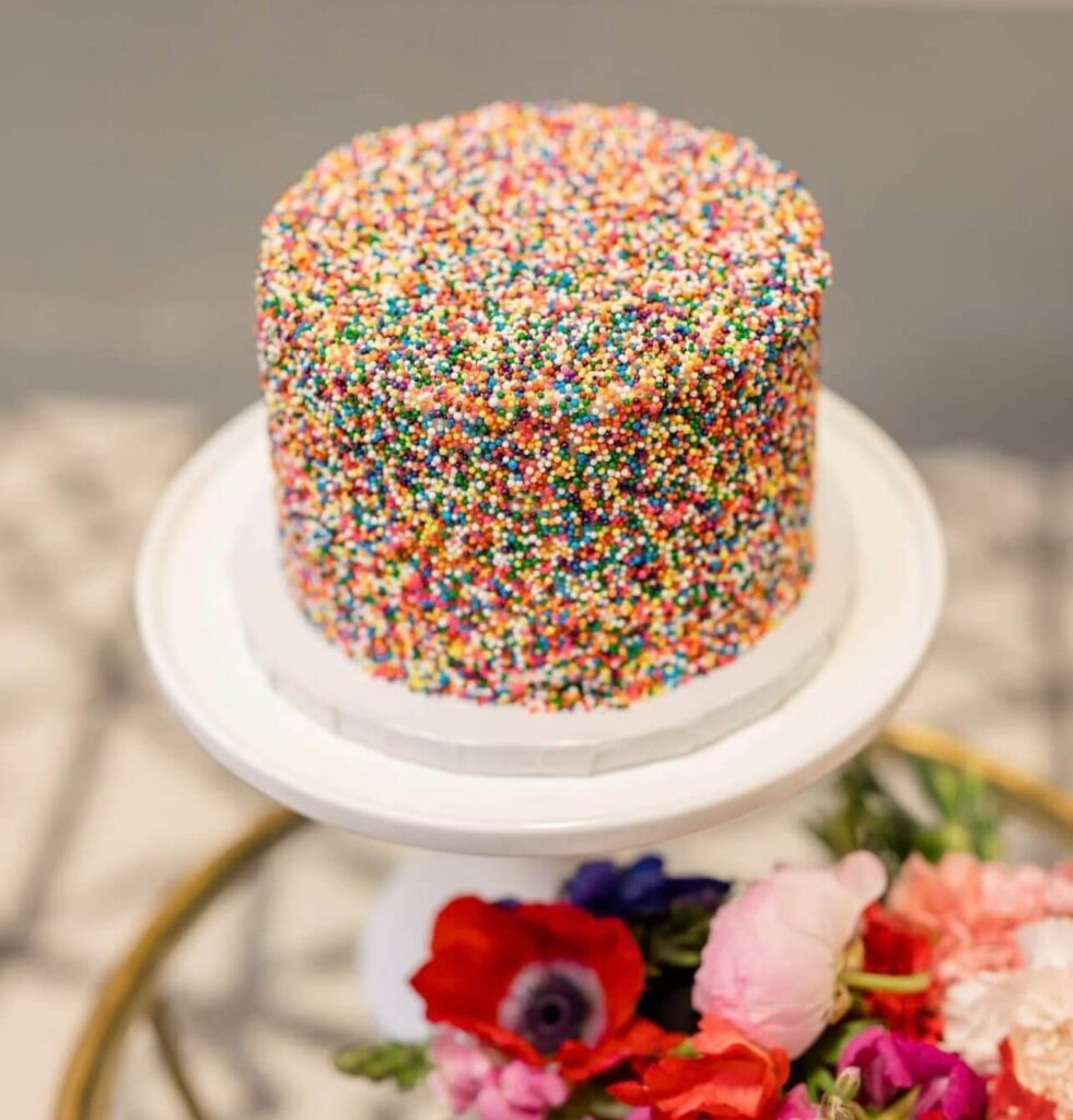 A cake completely covered in colorful rainbow nonpareil sprinkles, placed on a white cake stand.