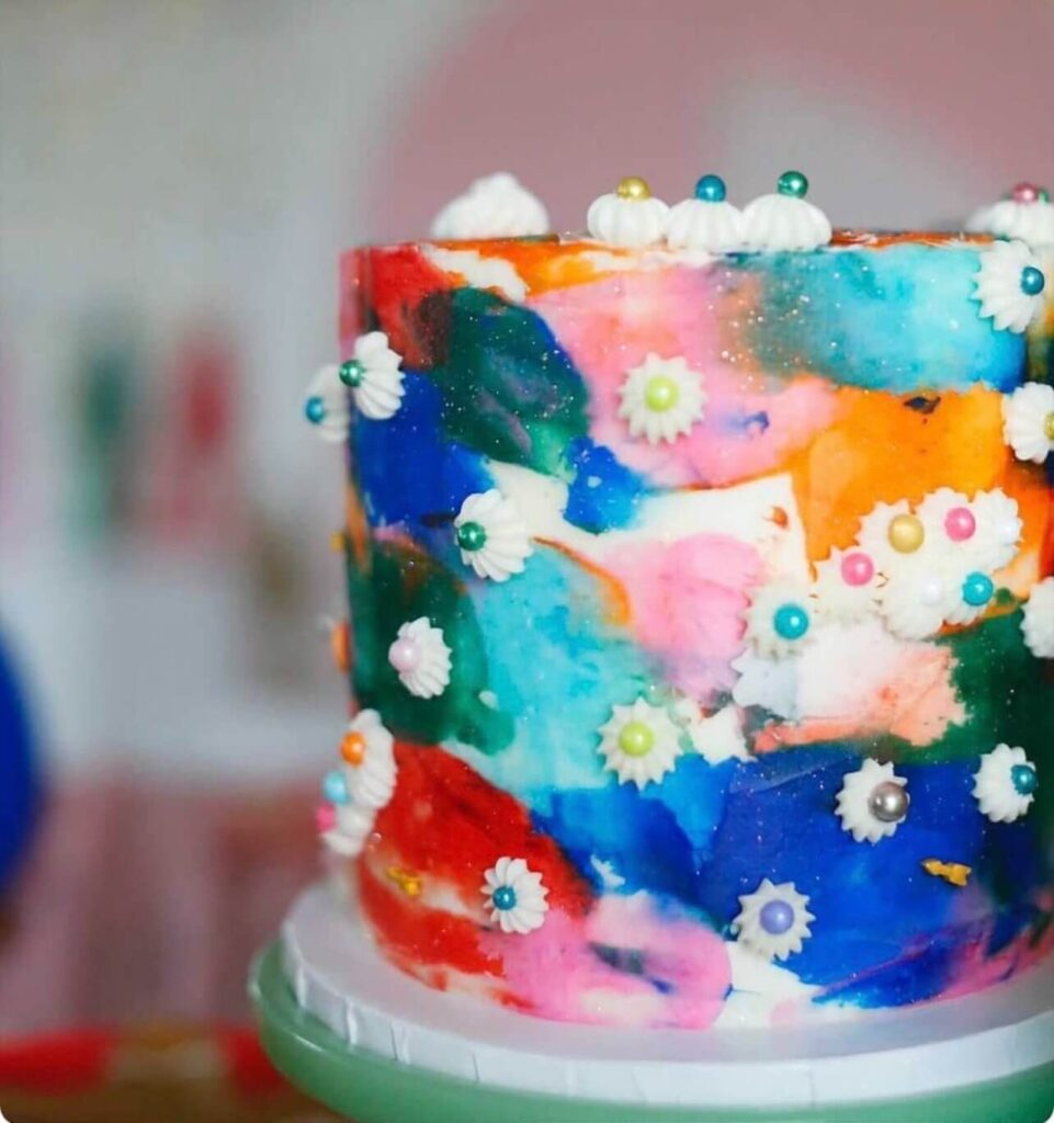 A painted buttercream cake in red, blue, and purple tones, topped with a bouquet of fresh roses.