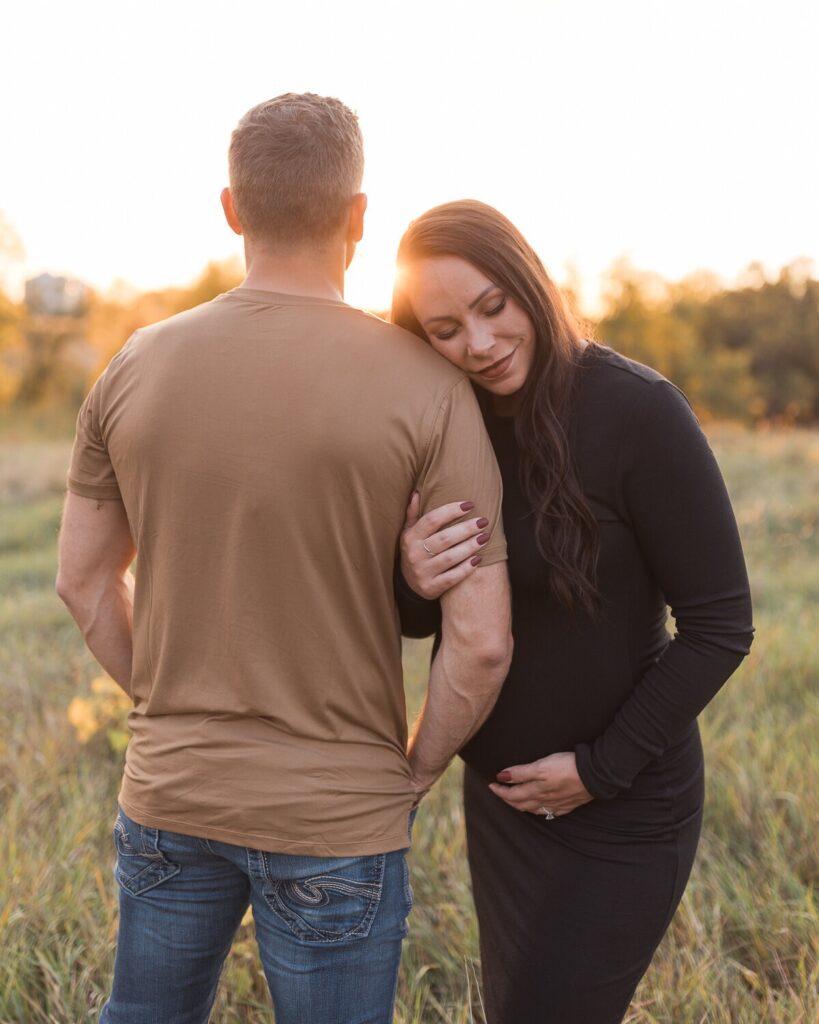 A pregnant woman rests her head on her partner’s shoulder in a field as she holds her belly and smiles softly at sunset.