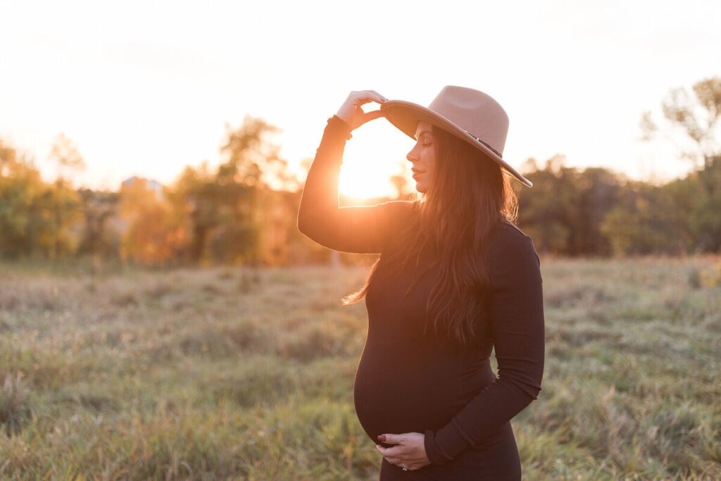 A pregnant woman in a black dress and wide-brimmed hat poses with one hand on her belly and the other resting on her head at sunset.