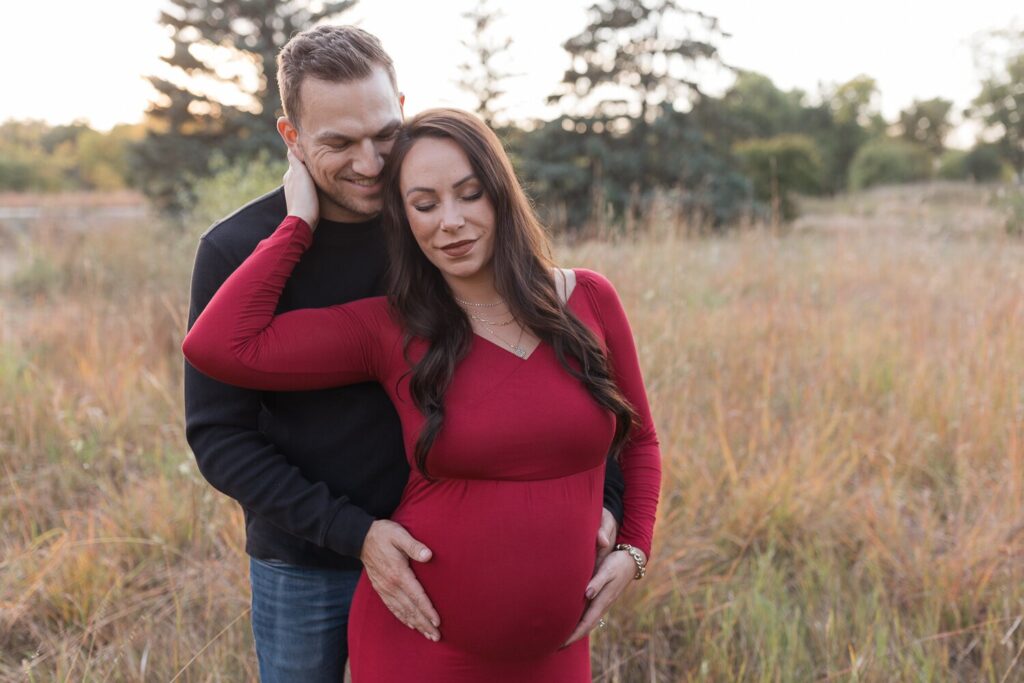 A couple stands in a field with the man smiling behind the pregnant woman in a red dress as they cradle her belly.