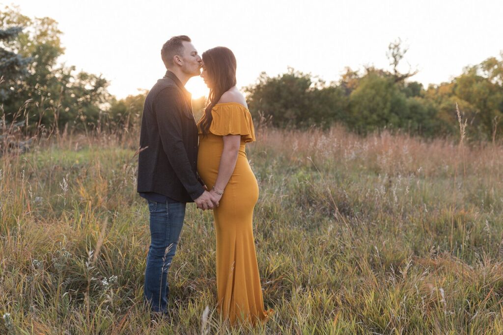 A couple stands in a grassy field at sunset, holding hands as the man kisses the pregnant woman’s forehead.