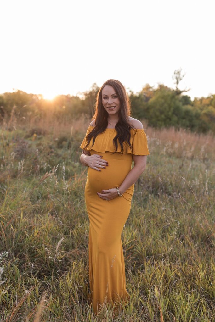 A smiling pregnant woman in a mustard yellow off-the-shoulder dress stands in a grassy field during golden hour.