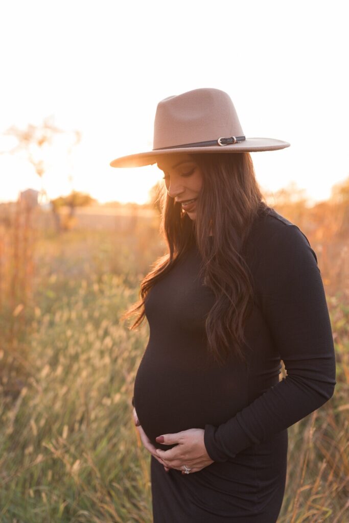 A maternity photo in a grassy field shows a woman in a black dress and hat holding her belly with the sun setting behind her.