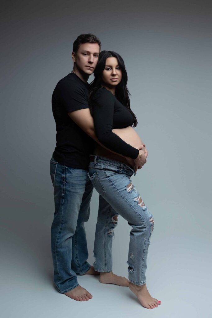 A couple stands barefoot in a studio, with the man embracing the pregnant woman from behind as she cradles her belly.