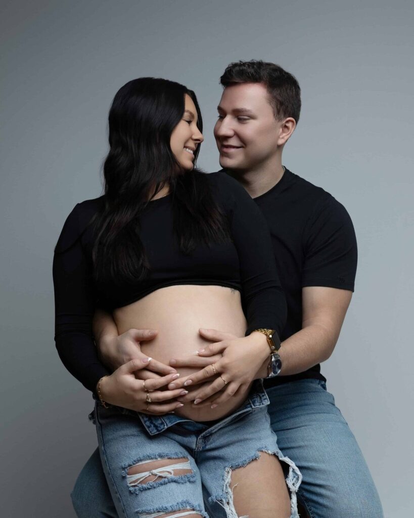A couple sits together in a studio, holding the woman's exposed pregnant belly and smiling lovingly at each other.