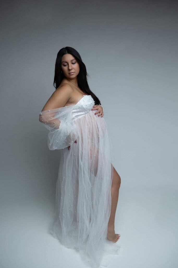 A maternity portrait of a woman in a sheer white robe, holding her belly and looking down gracefully in a studio setting.