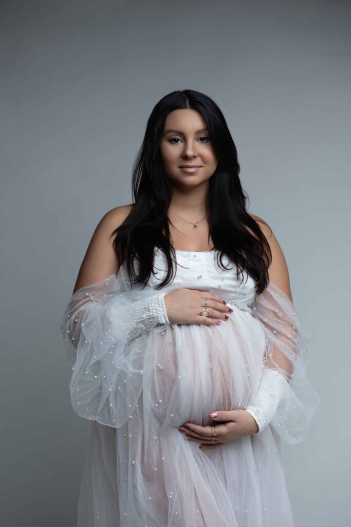A pregnant woman gently smiles while cradling her belly in a sheer white robe with pearl detailing.