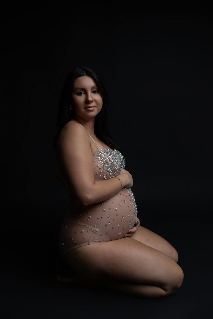 A woman kneels in a dark studio wearing a sheer bodysuit embellished with pearls, holding her belly and softly smiling.
