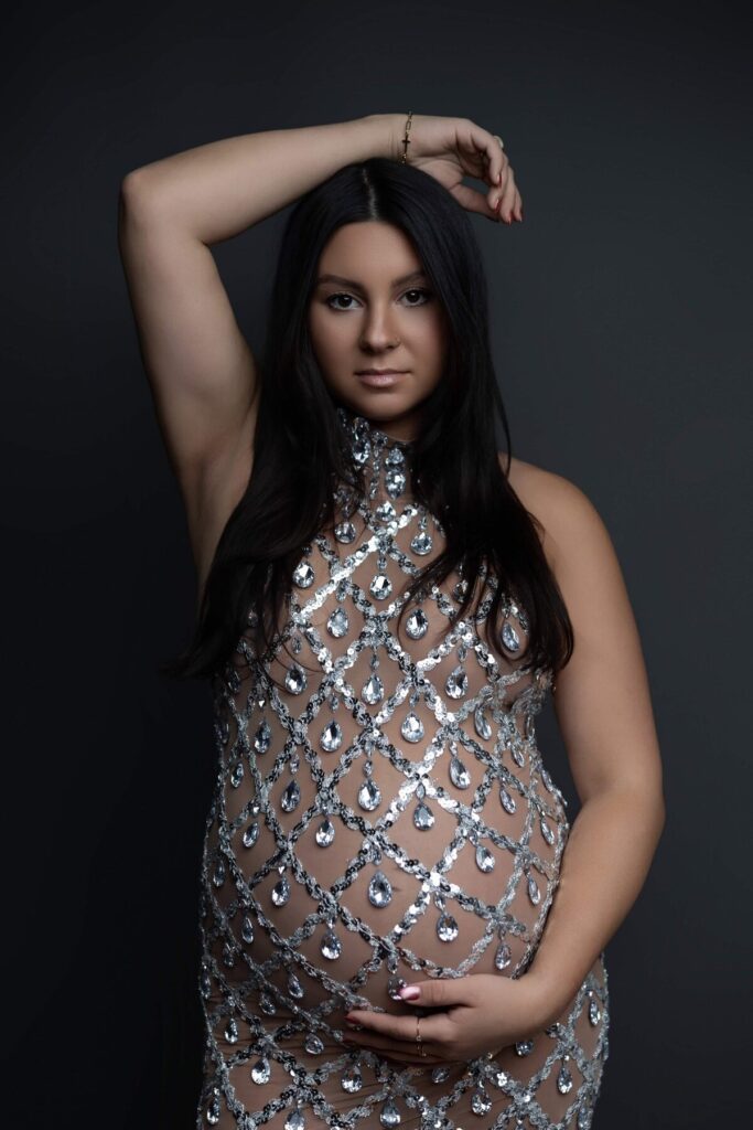 A pregnant woman poses in a glamorous sheer dress adorned with silver rhinestones, hand resting on her belly.