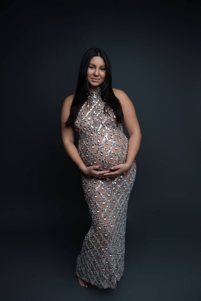 A full-length studio portrait of a pregnant woman in a glittering, gem-covered sheer gown, cradling her belly.