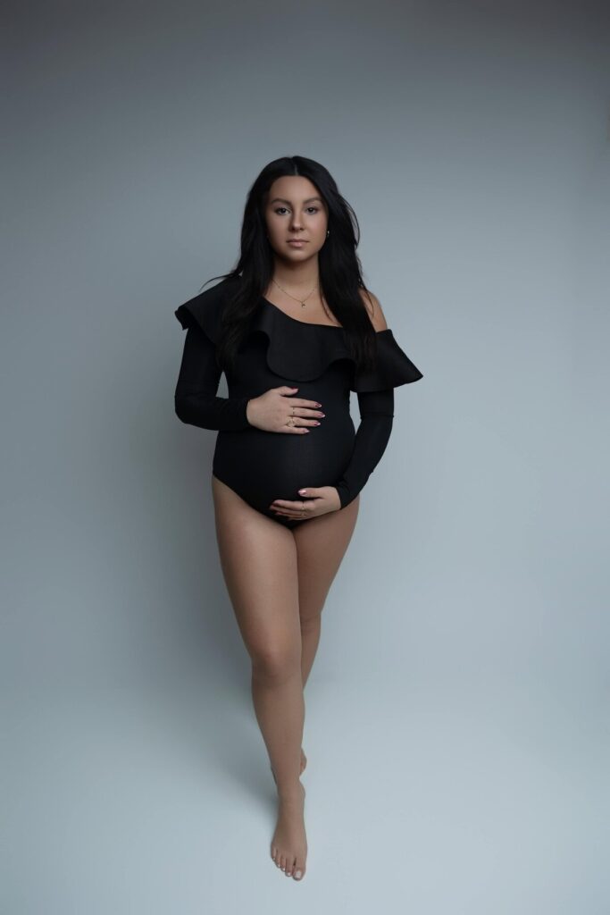 A standing maternity photo in a black off-shoulder bodysuit, with the woman softly holding her baby bump.