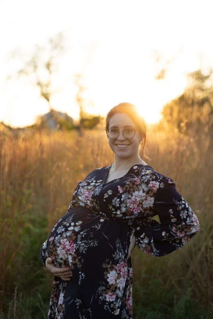 A pregnant woman in a floral dress smiles at the camera while holding her belly in a field at sunset.