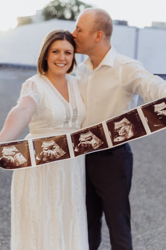 A smiling couple holds up ultrasound photos, with the man kissing the woman's forehead during their maternity shoot.
