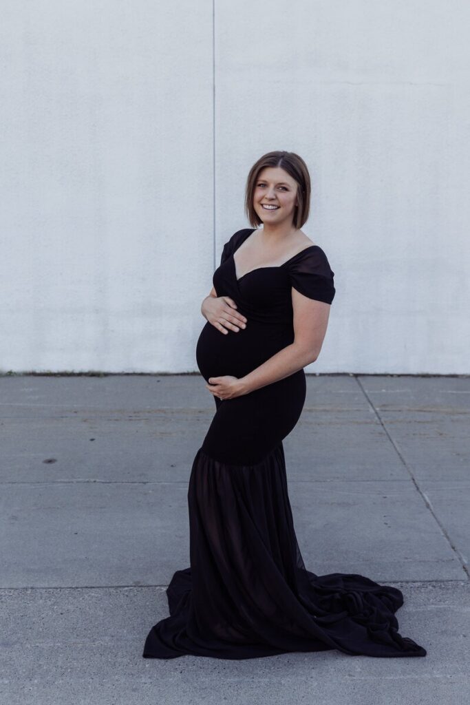 A pregnant woman in a fitted black gown poses outdoors against a light-colored wall, smiling and holding her belly.