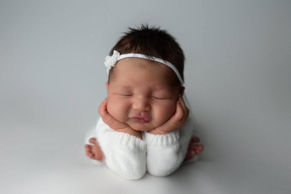 A newborn baby girl in a white outfit and headband sleeps with her hands propped under her chin, posed gently on a light grey studio backdrop.