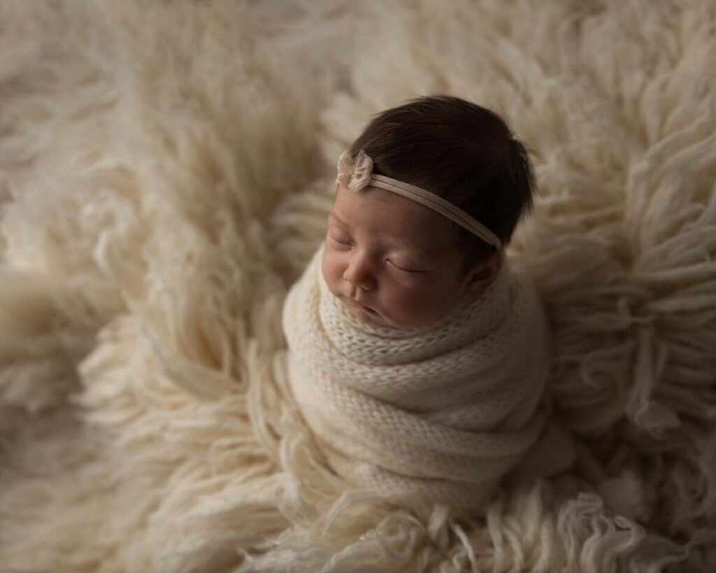 A newborn baby girl sleeps soundly while swaddled in a cream knit wrap, resting on a soft, fluffy textured blanket in a cozy studio setting.
