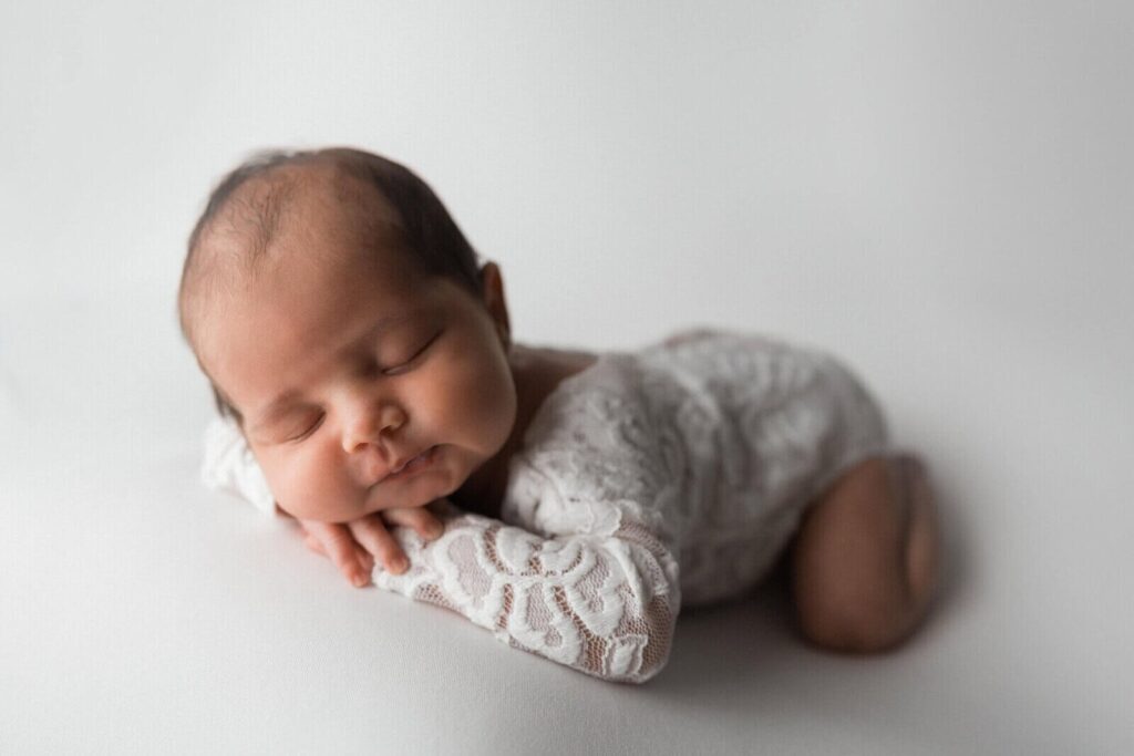 A newborn girl sleeps peacefully in a lace outfit on a white backdrop during a soft and elegant studio photoshoot.