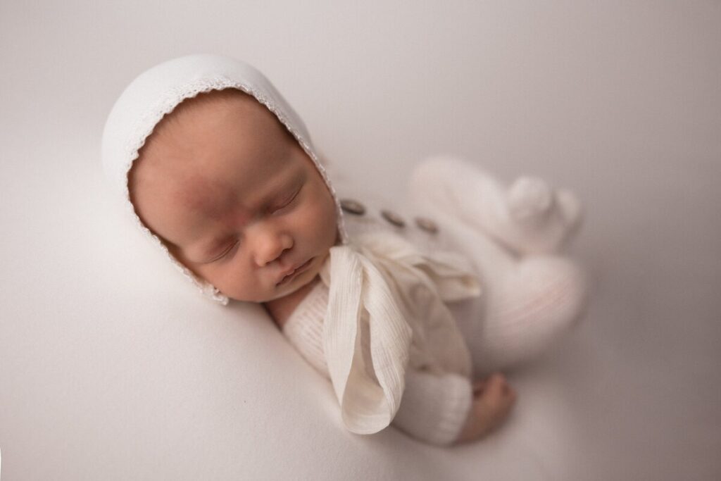 A newborn baby wearing a white bonnet and soft knit outfit sleeps on their side on a white backdrop in a studio setting.